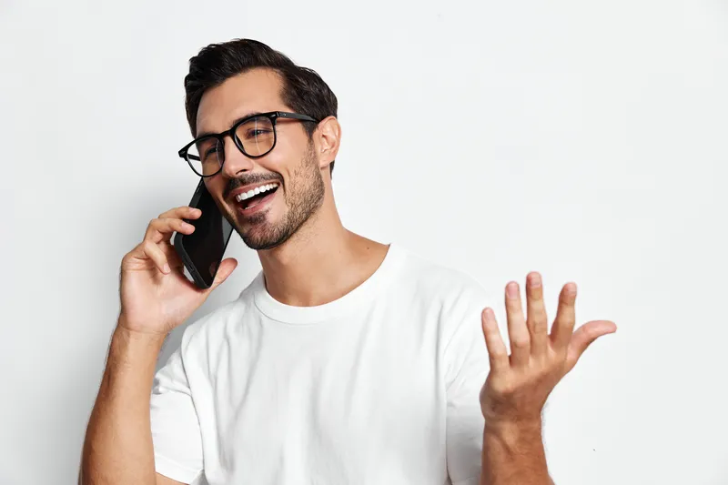 Smiling young man wearing glasses talking on mobile phone, expressing joy and excitement, isolated on plain solid white background. Communication lifestyle concept.