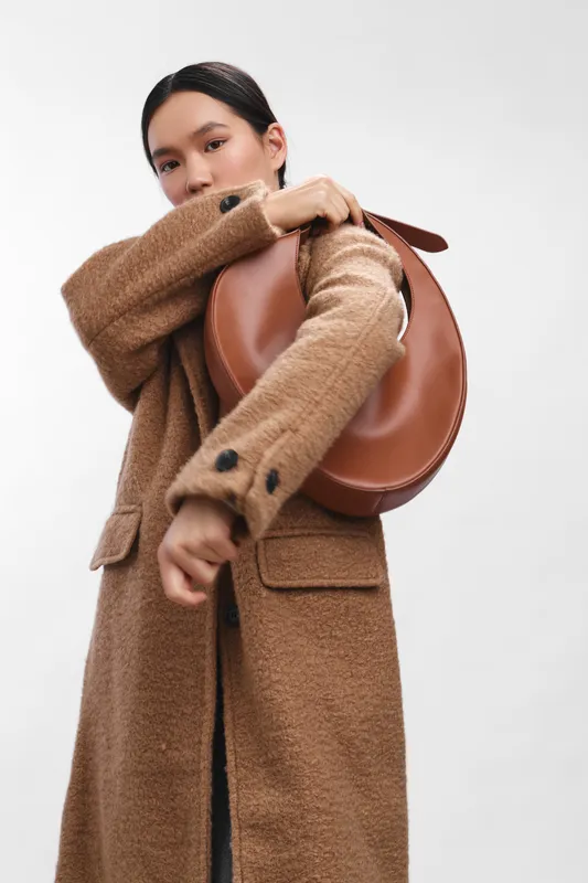 Asian woman wearing a warm tan coat with a round brown backpack, posed with a lifted sleeve, delivering a stylish and confident fashion moment in a clean studio setting with soft lighting and minimal