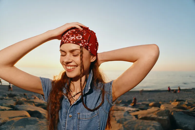 Lifestyle woman in modern boho-western denim street style smiling outdoors with a red bandana and film color effect, enjoying a sunny beach sunset moment.
