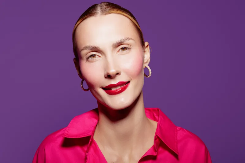Portrait of confident woman with red lipstick and hoop earrings wearing bright pink shirt against purple background. Concept of beauty, fashion, and self-expression.