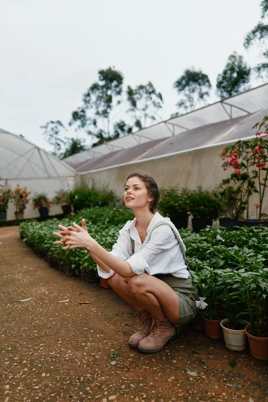 Young woman kneeling among vibrant greenery in a greenhouse, showcasing passion for gardening and nature, dressed in casual attire, expressing joy and connection