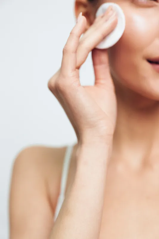 Young woman with clear skin gently applies a cotton pad to her cheek in a close-up beauty skincare routine on a light background, expressing calmness and self-care.