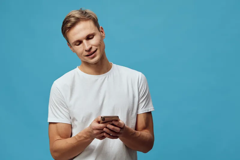 Smiling young man casual wear white t-shirt use mobile phone isolated on plain blue background studio portrait. People lifestyle concept