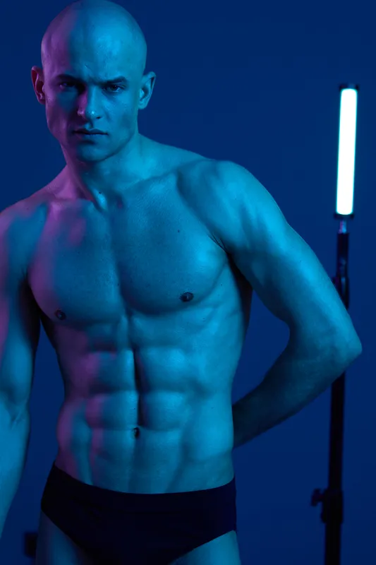 A muscular man poses dramatically under vibrant blue lighting, showcasing his toned physique. The striking color contrasts create a captivating aesthetic in this artistic representation.