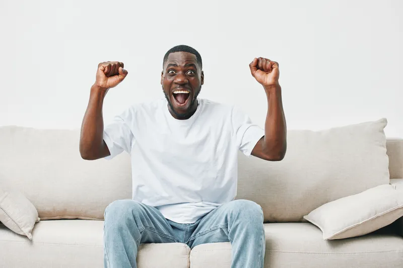 Excited young African man celebrating success on a light background with joyful expression, wearing white t shirt and jeans, perfect for uplifting themes