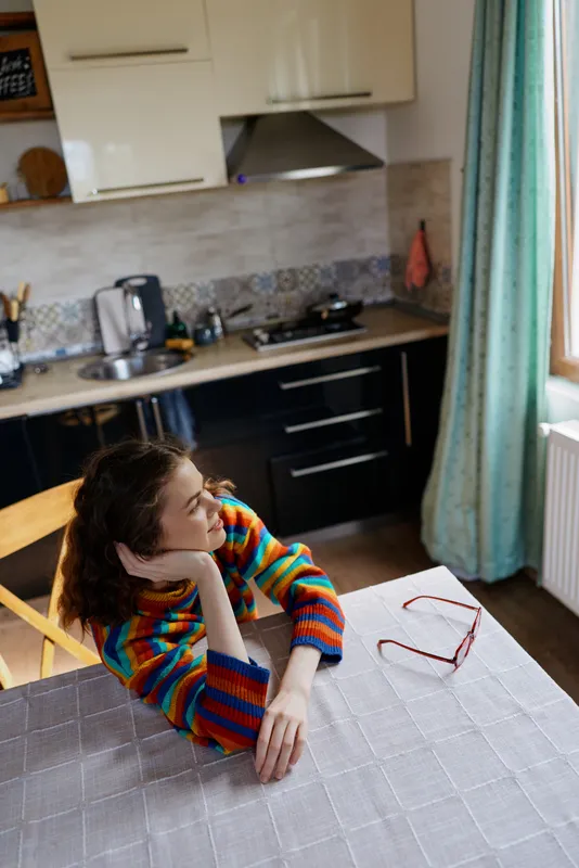 Contemplative woman in a colorful striped sweater rests her chin on her hand at a kitchen table, sunlight flowing through a window, a cozy home lifestyle moment