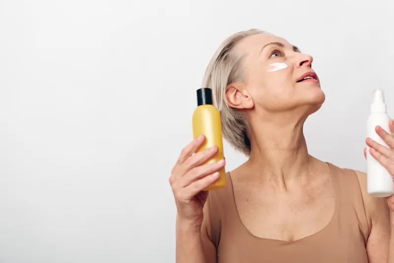 Mature woman with grey hair applies skincare cream on face while holding two cosmetic bottles. Concept of beauty routine, self-care, anti-aging, and healthy skin with natural products.