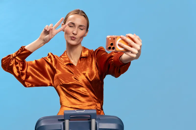 Young woman in shiny orange blouse takes selfie with peace sign while standing behind blue suitcase on plain background. Concept of travel, vacation, happiness, and modern lifestyle.