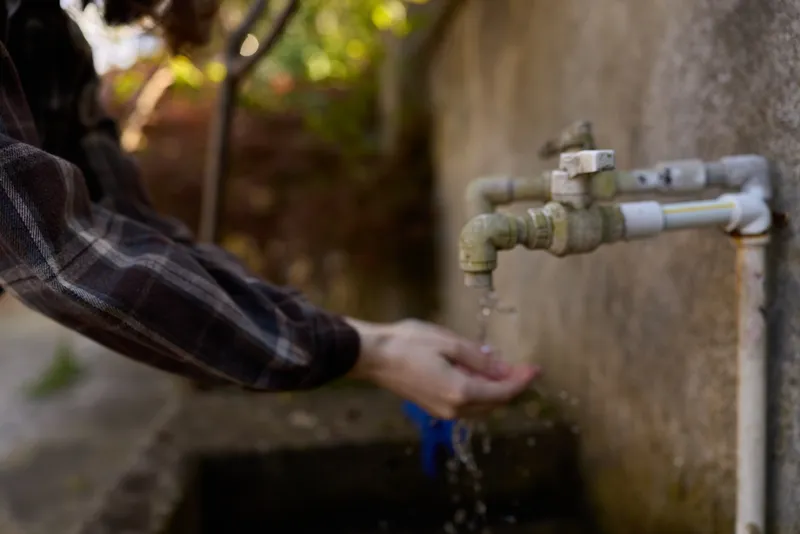 Candid outdoors scene of a woman washing hands at a rustic faucet, water droplets glint in sunlight, casual lifestyle moment, natural textures, simple everyday task
