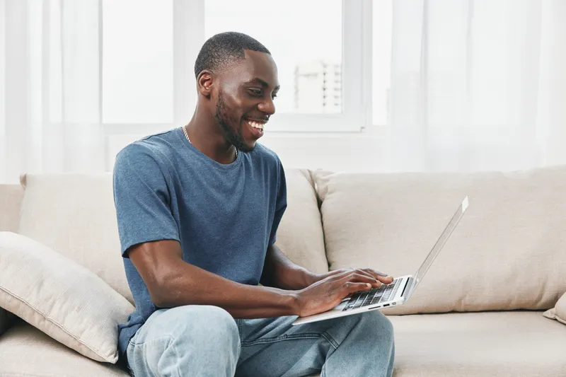 Young Black man smiling while using a laptop at home, dressed casually in a blue t shirt and jeans, showcasing a relaxed and happy lifestyle