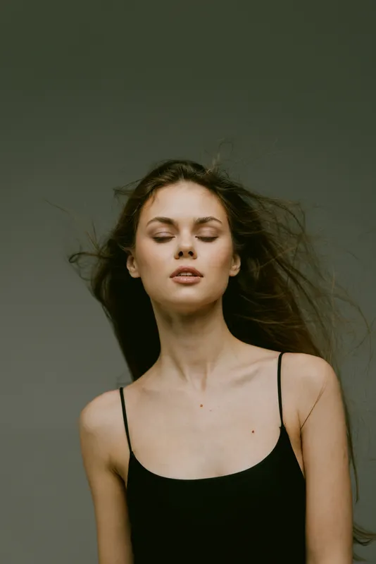 Young woman with closed eyes and flowing hair in black top expressing calm and serenity in soft lighting.