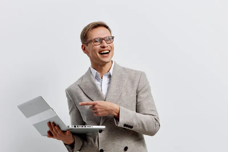 Happy middle-aged man with glasses in smart casuals holding laptop and pointing at it, smiling broadly, isolated on plain white background, studio portrait. People lifestyle concept
