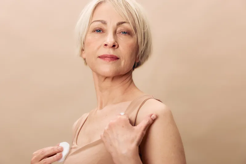 Mature woman with short blonde hair applies skincare cream on shoulder. Neutral background highlights natural beauty and self-care in a calm and soft lighting setting.