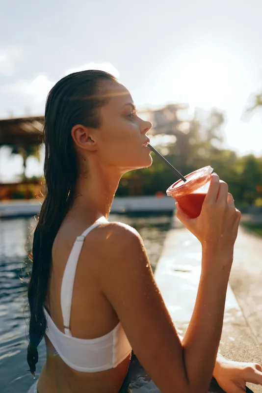 Sunlit poolside moment with a relaxed woman sipping a bright drink, wet hair, white swimsuit, casual leisure, vibrant colors, soft breeze, summer lifestyle, outdoor setting.