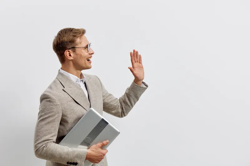 Businessman holding tablet and waving hand, smiling, wearing smart casual outfit, glasses, professional and confident expression, standing against plain light background, studio shot. People lifestyle