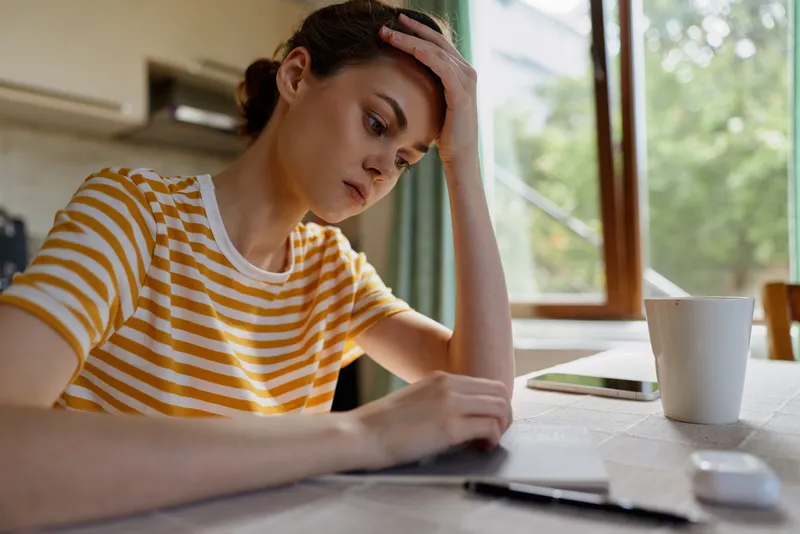 Woman at home work setup, resting her head on one hand, focused expression, striped shirt, notebook and laptop on table, coffee cup nearby, natural daylight and calm productive home mood