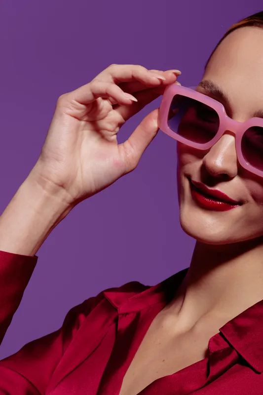 Stylish woman wearing pink sunglasses and red lipstick poses confidently against purple background. Fashion concept with vibrant colors and modern accessories for trendy look.