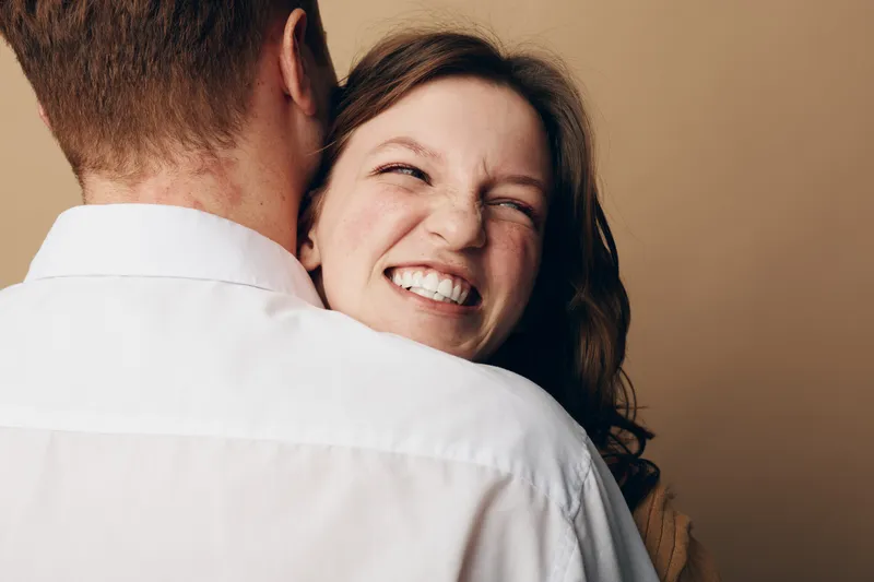Happy woman with curly hair smiles joyfully while hugging a man in white shirt. Close-up portrait with warm beige background. Concept of love, affection, and relationship happiness.
