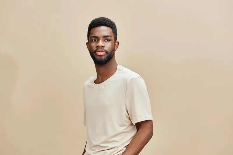 Young African man in casual beige outfit standing confidently against a neutral background, showcasing a relaxed yet thoughtful expression