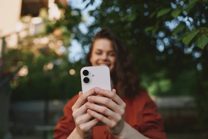 Cheerful woman outdoors taking a selfie, smiling behind a smartphone, sunlit moment with natural colors, casual jacket, greenery backdrop, lifestyle portrait vibe in ambient breeze