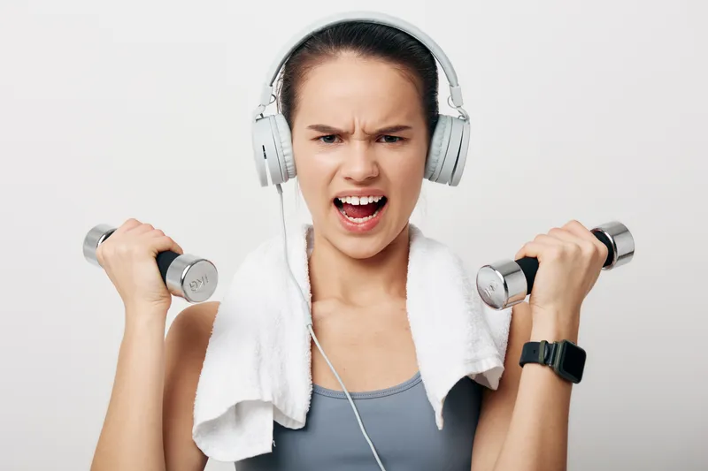 Woman fitness enthusiast lifting dumbbells in a gym, wearing headphones and a towel, showing energy and determination during an intense workout in a bright studio. She stays focused on form as she