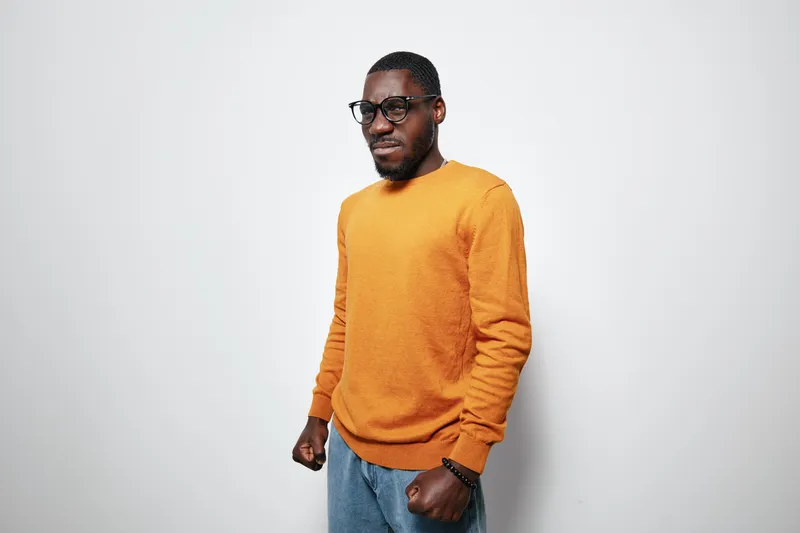 African man with dark skin wearing glasses and orange sweater standing with clenched fists on white background. Confident male portrait with casual style and facial expression.