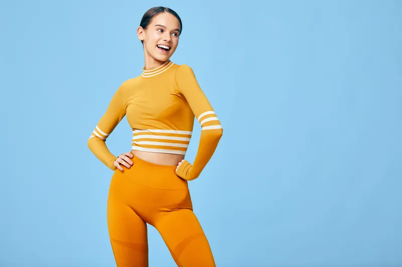 Fitness woman in orange athletic set strikes a confident pose against a blue studio backdrop, showcasing sporty attire, active lifestyle and motivation for workouts.