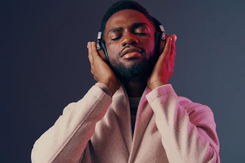 Young Black man enjoying music with headphones, wearing a stylish light coat, set against a gradient background with soft pink and purple tones, conveying relaxation and joy