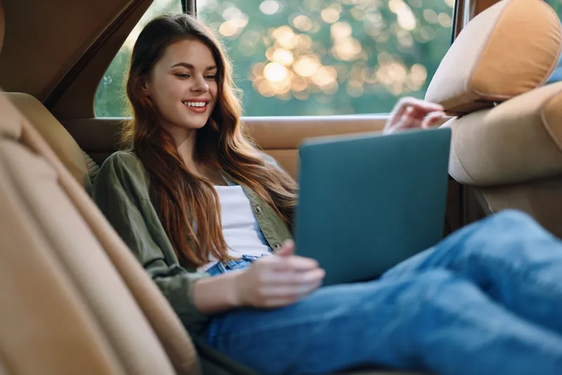 Young woman with long hair working on a laptop inside a car during sunset, wearing a casual outfit, smiling happily, immersed in her tasks.
