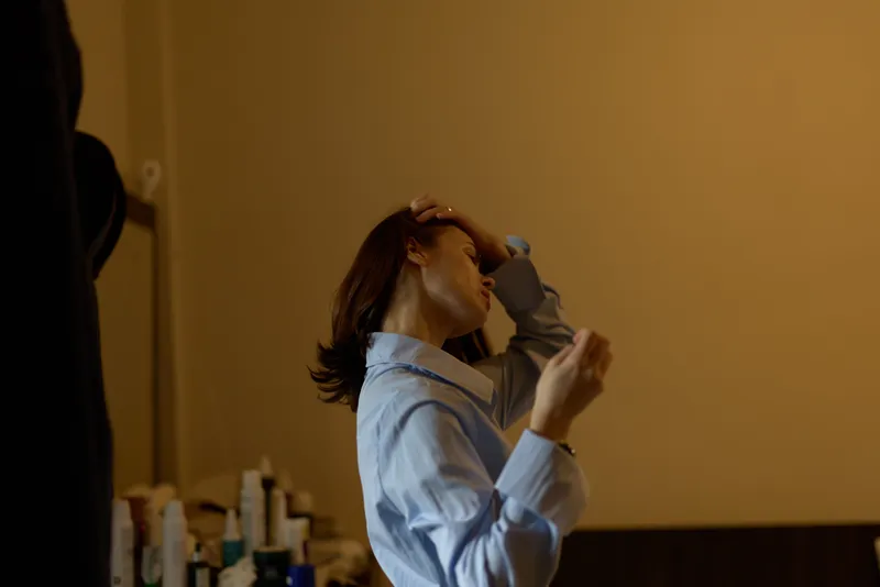 A woman in a blue shirt adjusts her hair while standing indoors near a table with bottles in a softly lit room, capturing a quiet personal moment.