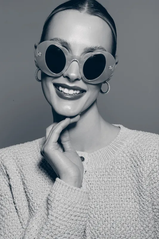 Smiling Caucasian woman with pale skin wears large round sunglasses and knitted sweater, posing with hand near chin in studio.