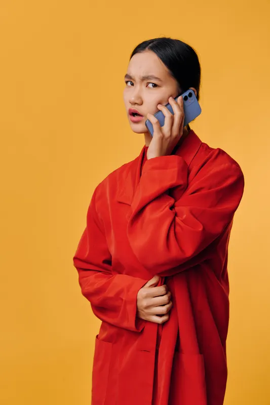 Woman in red coat holding smartphone near ear with confused expression, standing in front of orange background. Female talking on mobile phone, communication and emotion concept in studio.
