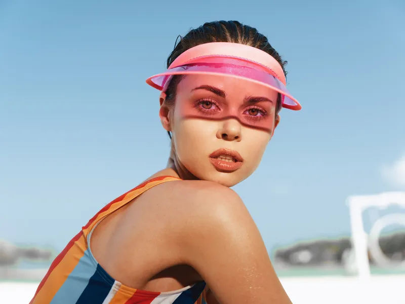 Portrait of a young woman at the beach wearing a pink visor and striped swimsuit, sunlit skin, and a confident gaze, capturing a bright summer moment.