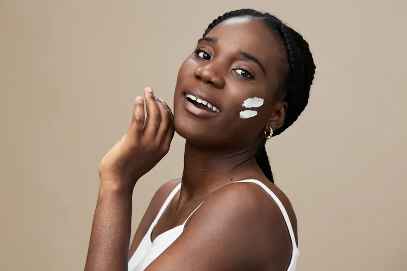 A joyful woman applying skincare with a smooth cream on her cheek, showcasing natural beauty and self care in a minimalist setting