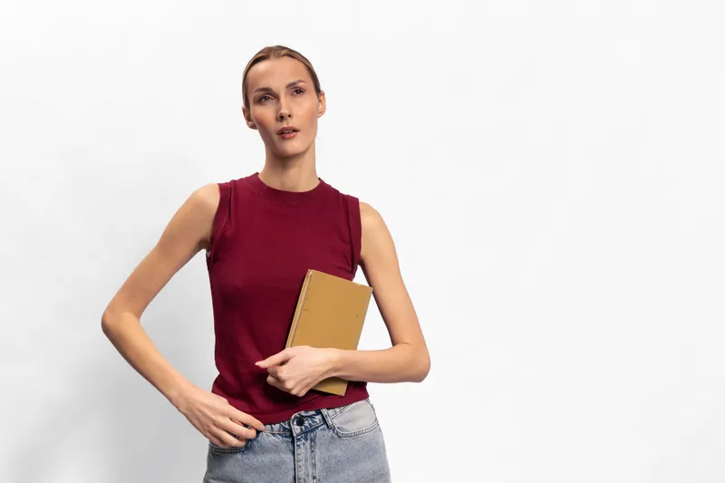 Young woman in sleeveless red top holds notebook, standing against white background. Casual style with blue jeans. Concept of education, study, planning, and modern lifestyle.