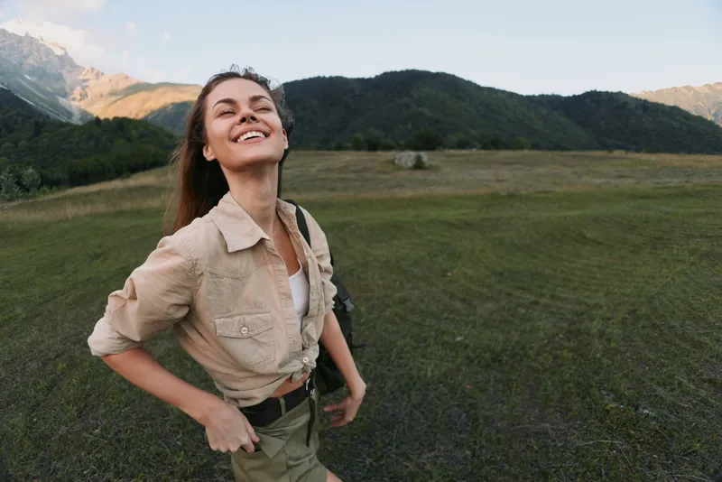 Happy woman enjoys outdoor lifestyle in nature with mountains and green hills behind, smiling with closed eyes, wearing casual beige shirt and cargo pants.