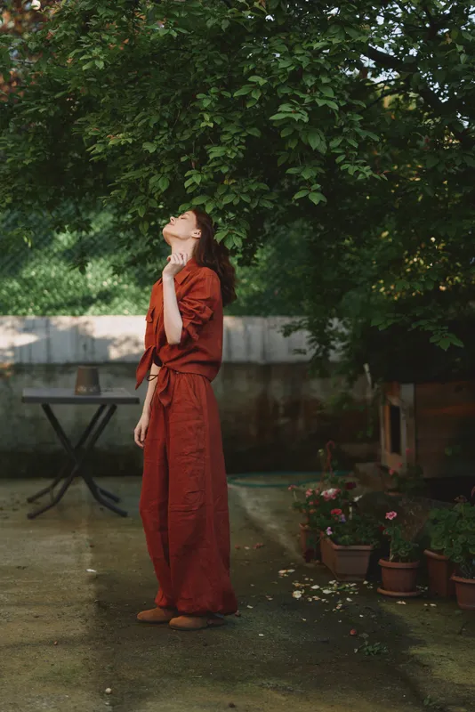 A woman in a rust red dress stands outdoors, head tilted up toward the sky, in a sunlit garden. A calm, reflective moment in everyday life