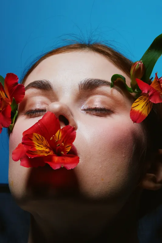 Female model with closed eyes, surrounded by vibrant red flowers on a blue background, expressing tranquility and beauty in a fresh, creative concept