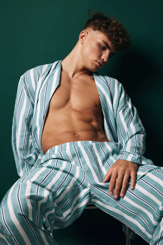 Young man with curly hair wearing striped pajamas sitting on chair with relaxed pose against dark background. Casual fashion, male model and comfortable homewear style.