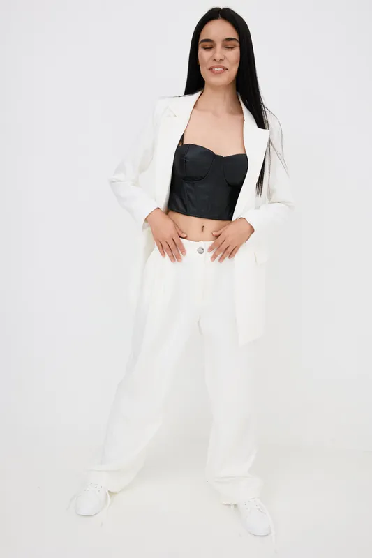 Young woman dressed in stylish white suit with black corset, posing confidently with eyes closed against a plain white background in modern fashion style.
