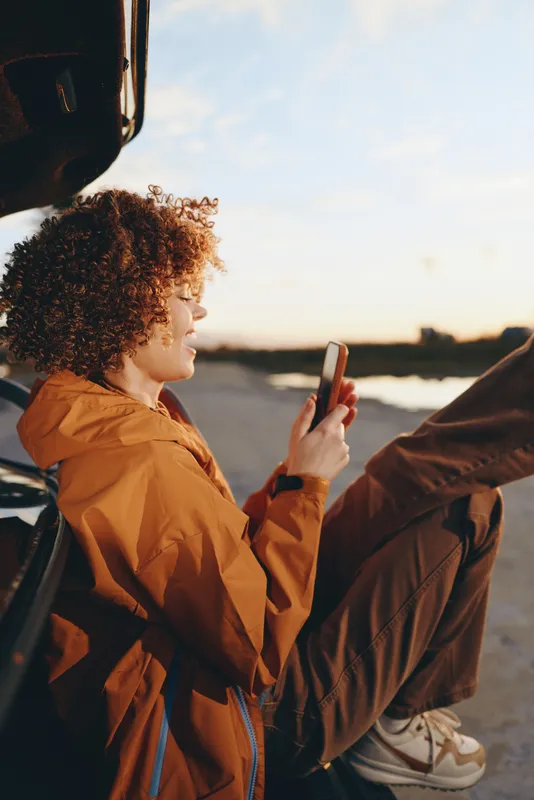 Woman lifestyle outdoor sunset, sitting in a car, holding a phone, wearing an orange jacket, curly hair in profile, warm evening light, relaxed trip feeling, by the road at golden hour