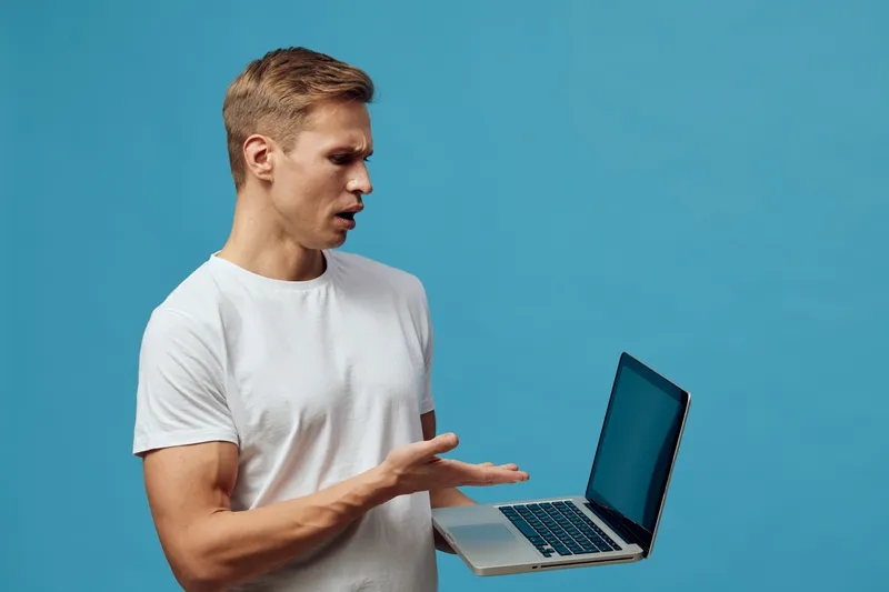 Man holding open laptop computer in hand, looking at screen with focused expression, casual white t-shirt, blue background, studio portrait, technology, work, digital device, communication, modern, pr