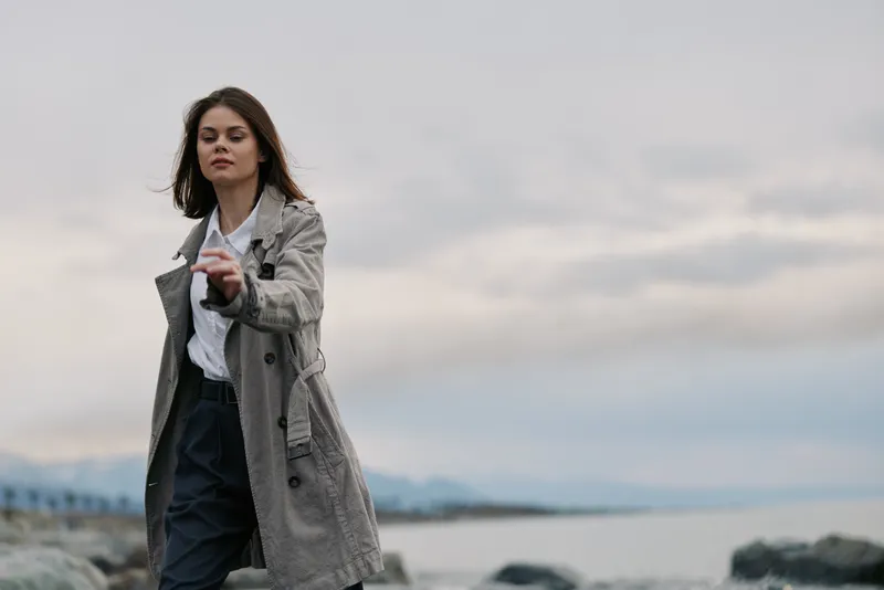 Young woman in a long coat walks outdoors near rocky shore with cloudy sky. Casual fashion, autumn mood, and calm natural environment with sea and distant mountains in background.