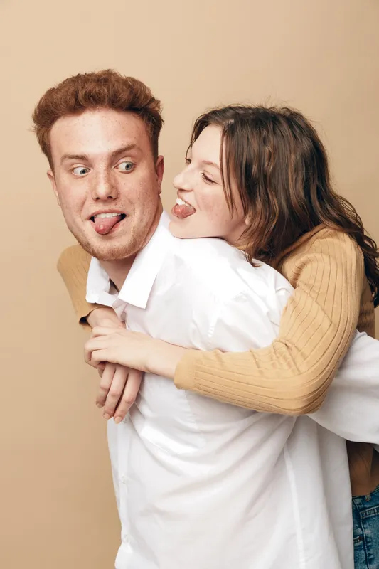 Playful young couple with red hair and brown hair making funny faces, woman hugging man from behind. Casual clothing and beige background create a warm, joyful mood of friendship and fun.