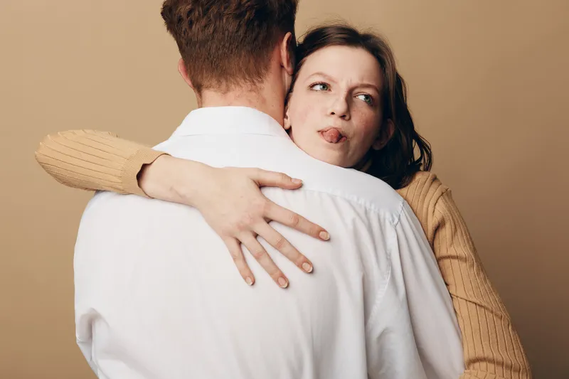 A woman in a beige sweater hugs a man in a white shirt but looks uncomfortable and distant. The image shows complex emotions in a close relationship, concept of tension and doubt.