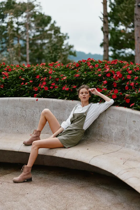 A fashionable young woman in a stylish olive green overall dress poses gracefully outdoors among vibrant flowers, showcasing a relaxed and confident expression
