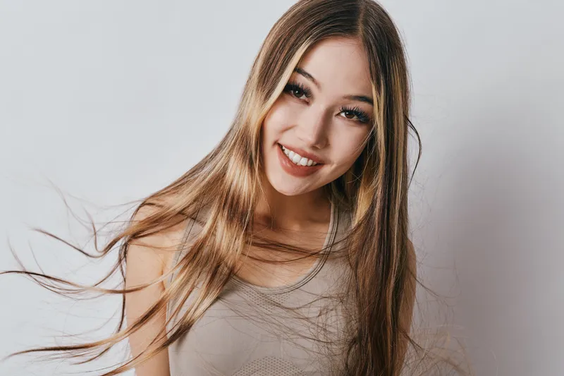 Young woman with long hair smiling at camera with natural makeup against plain background in casual outfit for beauty and lifestyle portrait.