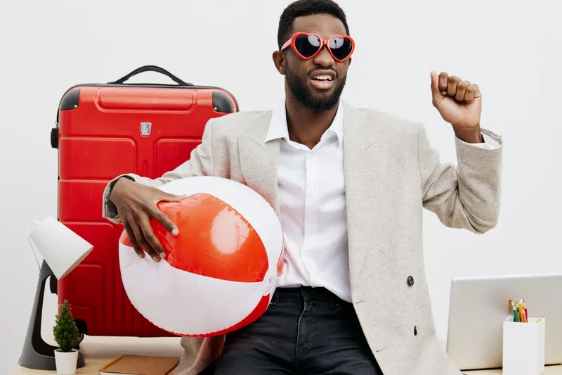 Young smiling man with dark skin, wearing beige blazer, holding an orange and white beach ball, sitting at desk with travel suitcase, computer and stationery on white background. People lifestyle conc