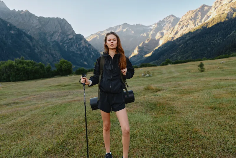 A lone female hiker stands in a wide mountain meadow with trekking pole and camera gear, ready for an outdoor adventure. She wears a dark jacket and shorts, framed by rugged peaks, creating a calm,