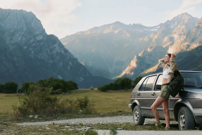 Woman leaning on a car in a mountain landscape, casual outfit, travel lifestyle, outdoor adventure, nature and serene mood, warm sunlight and expansive horizon.